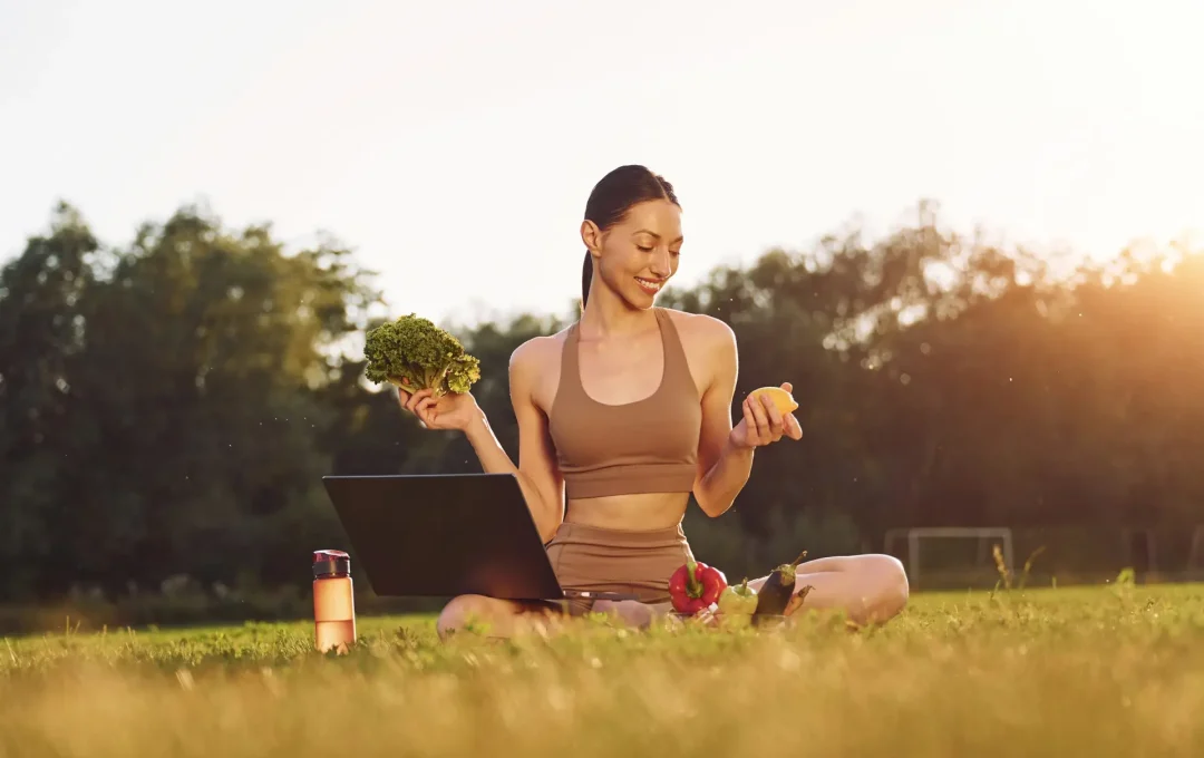 Using laptop. Holding vegetables. Young woman in yoga clothes is outdoors on the field.